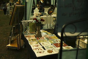 A market stall displays an array of vibrant greeting cards and floral arrangements. Next to the cards, a decorative sign and small ornaments are placed on the table. In the background, people browse through other stalls, creating a lively and community-focused atmosphere.