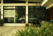 A reflective glass window displays the interior of a room with a parked silver car. The exterior view includes a concrete pavement, some plants, and a few chairs. Palm trees are faintly visible through the reflection.