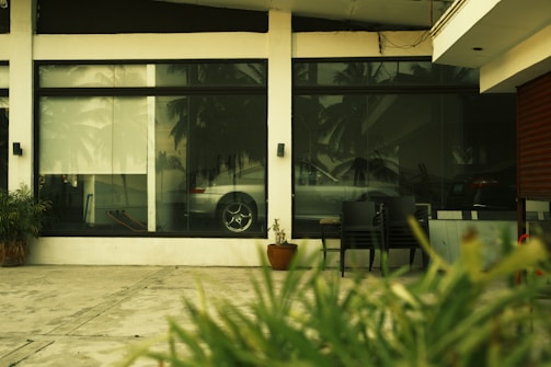 A reflective glass window displays the interior of a room with a parked silver car. The exterior view includes a concrete pavement, some plants, and a few chairs. Palm trees are faintly visible through the reflection.