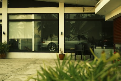 A reflective glass window displays the interior of a room with a parked silver car. The exterior view includes a concrete pavement, some plants, and a few chairs. Palm trees are faintly visible through the reflection.