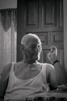 An elderly person with short hair sits in front of a closed wooden door, wearing a sleeveless shirt, and appears to be making a gesture with one hand. The setting suggests an indoor environment with faint natural light coming through a window, partially covered with lace curtains.