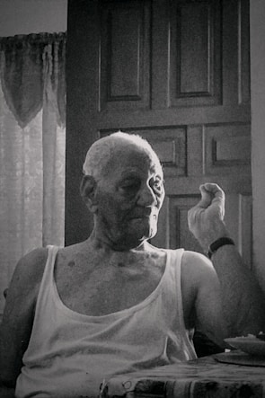 An elderly person with short hair sits in front of a closed wooden door, wearing a sleeveless shirt, and appears to be making a gesture with one hand. The setting suggests an indoor environment with faint natural light coming through a window, partially covered with lace curtains.