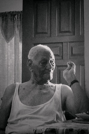 An elderly person with short hair sits in front of a closed wooden door, wearing a sleeveless shirt, and appears to be making a gesture with one hand. The setting suggests an indoor environment with faint natural light coming through a window, partially covered with lace curtains.
