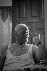 An elderly person with short hair sits in front of a closed wooden door, wearing a sleeveless shirt, and appears to be making a gesture with one hand. The setting suggests an indoor environment with faint natural light coming through a window, partially covered with lace curtains.