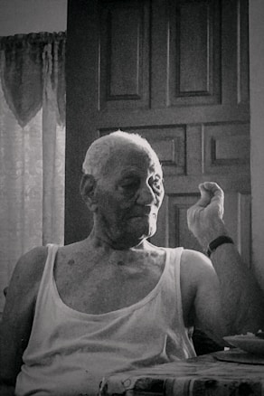 An elderly person with short hair sits in front of a closed wooden door, wearing a sleeveless shirt, and appears to be making a gesture with one hand. The setting suggests an indoor environment with faint natural light coming through a window, partially covered with lace curtains.
