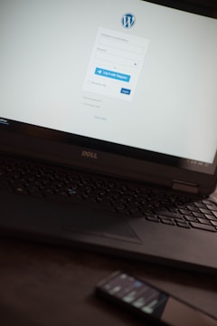 A laptop displaying a WordPress login screen with fields for username and password. The screen has options to log in with Telegram or email. The logo of WordPress is visible on top. A smartphone is resting on the surface near the laptop.