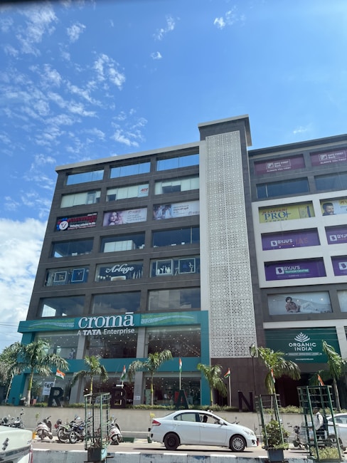 A multi-story commercial building with various advertisements and brand signage on its facade. The ground floor has a Croma store, and above it are signs for different brands and services. Palm trees are planted along the sidewalk, and several vehicles, including a white car and scooters, are parked or moving in front of the building. The sky is mostly clear with scattered clouds.