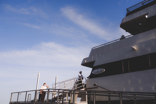 A large cruise ship is docked, featuring a person in uniform standing on the deck near the railing under a clear blue sky. The ship is named 'Odyssey' with its name visible on the side. The scene conveys an atmosphere of anticipation or readiness.