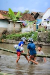 Two children wearing helmets are playing on a bamboo structure above a shallow body of water. They are holding onto each other and seem to be balancing on the bamboo. Another person is in the water nearby, watching or assisting them. In the background, there are houses with tiled roofs and large banana plants.