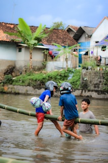 Two children wearing helmets are playing on a bamboo structure above a shallow body of water. They are holding onto each other and seem to be balancing on the bamboo. Another person is in the water nearby, watching or assisting them. In the background, there are houses with tiled roofs and large banana plants.