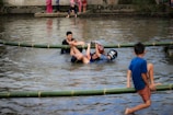 Several people are playfully engaged in a game involving climbing or balancing on bamboo poles placed over a shallow body of water. One person, wearing a helmet, is grabbing onto another participant, while a third person in blue shorts is visibly enjoying himself. Spectators in bright clothing stand by the water's edge.