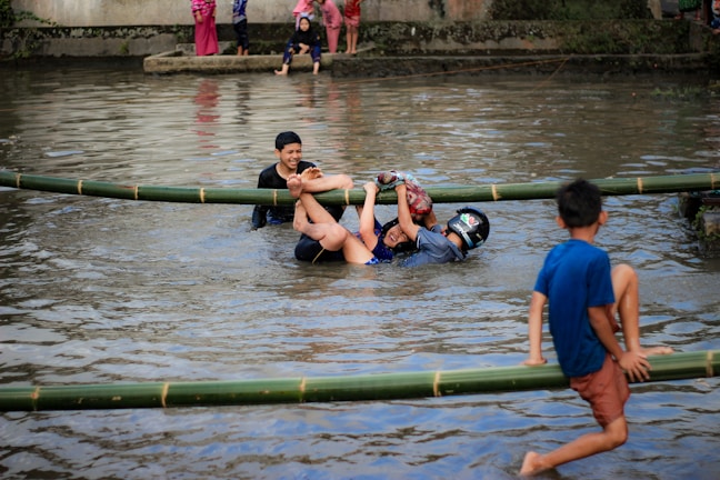 Several people are playfully engaged in a game involving climbing or balancing on bamboo poles placed over a shallow body of water. One person, wearing a helmet, is grabbing onto another participant, while a third person in blue shorts is visibly enjoying himself. Spectators in bright clothing stand by the water's edge.