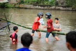 A group of young people are engaging in a playful activity in a body of water. Two individuals are balancing on a bamboo beam while wearing helmets, appearing to participate in a friendly competition. They seem to be trying to knock each other off while holding pillows. Other people are watching and standing in the shallow water, enjoying the game.