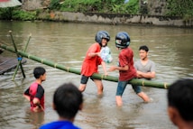 A group of young people are engaging in a playful activity in a body of water. Two individuals are balancing on a bamboo beam while wearing helmets, appearing to participate in a friendly competition. They seem to be trying to knock each other off while holding pillows. Other people are watching and standing in the shallow water, enjoying the game.