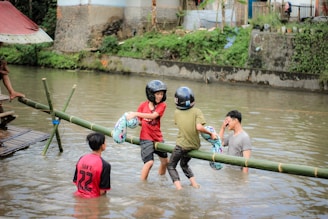Several individuals are playing in a river, where two of them balance on a bamboo pole above the water. They wear helmets and hold inflatable rings, and others stand in the river. A building and greenery are visible in the background.