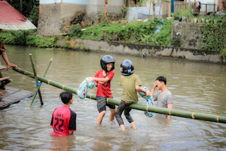 Several individuals are playing in a river, where two of them balance on a bamboo pole above the water. They wear helmets and hold inflatable rings, and others stand in the river. A building and greenery are visible in the background.