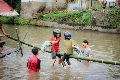 Several individuals are playing in a river, where two of them balance on a bamboo pole above the water. They wear helmets and hold inflatable rings, and others stand in the river. A building and greenery are visible in the background.