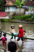 Children are playing in a body of water, balancing on a bamboo log while wearing helmets and holding colorful pillows. A man is in the water, supporting or observing them, with a house and greenery in the background.