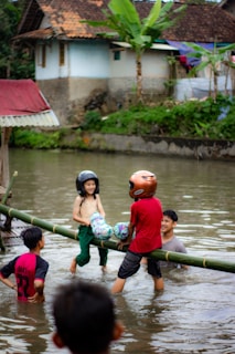 Children are playing in a body of water, balancing on a bamboo log while wearing helmets and holding colorful pillows. A man is in the water, supporting or observing them, with a house and greenery in the background.