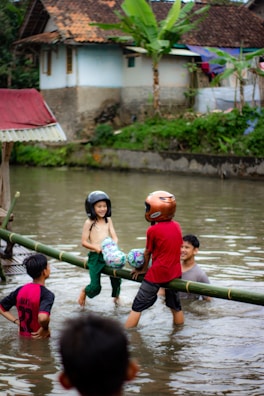 Children are playing in a body of water, balancing on a bamboo log while wearing helmets and holding colorful pillows. A man is in the water, supporting or observing them, with a house and greenery in the background.