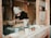 A person wearing a patterned headscarf and mask is working on crafting pottery on a wheel inside a workshop. Various ceramic mugs and cups are displayed on the windowsill in front of them. The workshop has a wooden interior, and the person appears focused on shaping the clay.