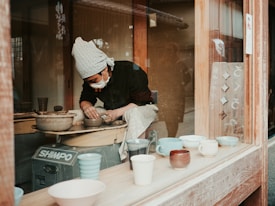 A person wearing a patterned headscarf and mask is working on crafting pottery on a wheel inside a workshop. Various ceramic mugs and cups are displayed on the windowsill in front of them. The workshop has a wooden interior, and the person appears focused on shaping the clay.
