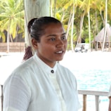 An individual wearing a white shirt stands in a tropical outdoor setting. Behind her are lush palm trees, a sandy beach, and a glimpse of turquoise water. A small vehicle is visible in the background next to a thatched structure.