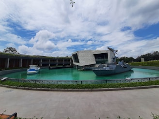 A modern architectural building with a grey facade is situated by a large artificial water feature, in which a navy ship is docked. The sky above is partly cloudy with scattered patches of blue. There is a smaller boat also visible on the left side of the water, surrounded by a low railing and greenery.
