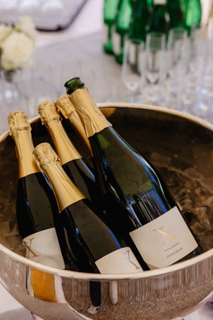 Several bottles of champagne are placed in a large metal bowl filled with ice. The bottles have gold foil on the neck and labels with a prominent 'X'. In the background, there are green glass bottles and empty champagne flutes, suggesting a celebratory setting.