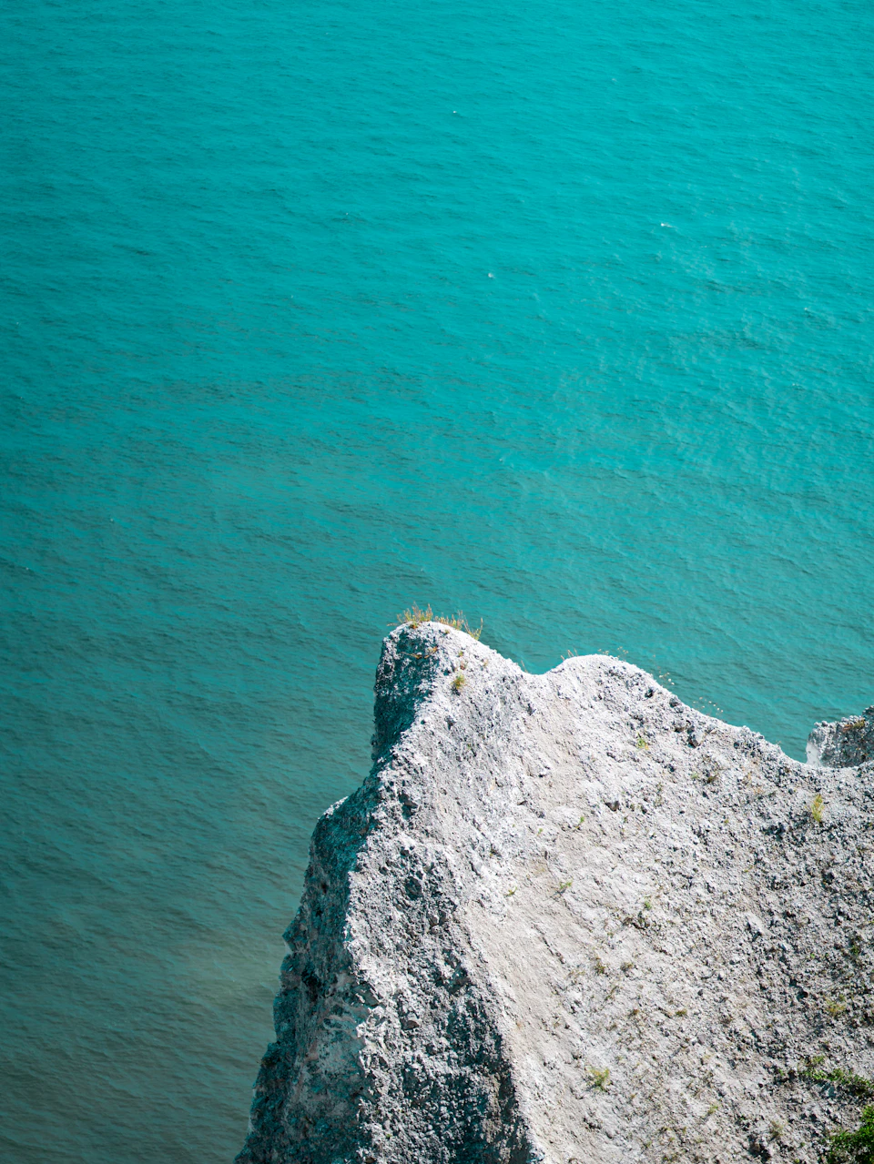 The image depicts a cliff overlooking a body of water, possibly an ocean or sea. The cliff is made of gray and white rocks that are jagged and unevenly shaped, giving it a rugged appearance. The water below has a light blue color, suggesting a calm and serene environment. The perspective of the photo is from above, providing a bird's eye view of the cliff and its surroundings.