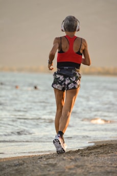 A person with short hair wearing a red sports top and floral shorts is jogging along a sandy beach. They are wearing headphones and athletic shoes. In the background, a body of water is visible with a few people swimming in the distance.