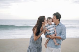 A family stands together on a beach, with a woman on the left kissing a baby held by a man on the right. The ocean and sky are visible in the background, creating a serene setting.