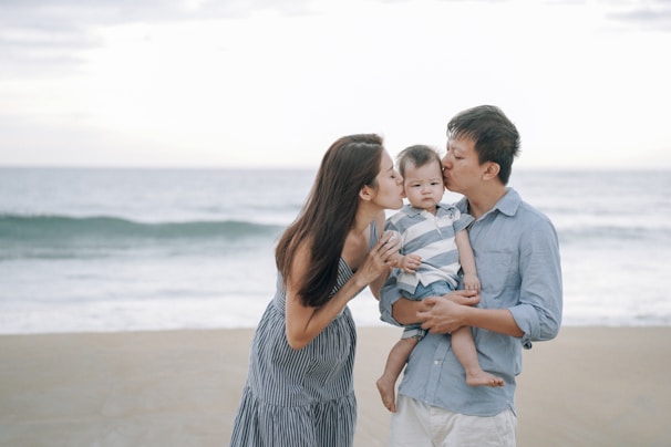 A family stands together on a beach, with a woman on the left kissing a baby held by a man on the right. The ocean and sky are visible in the background, creating a serene setting.
