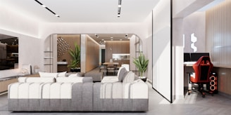 A contemporary living space featuring a large gray sectional sofa, an adjacent area with a gaming setup and ergonomic red and black chair, and a dining area in the background with a minimalist design. The room is well-lit with modern recessed lighting and contains decorative plants and sleek furniture. The walls and flooring are in neutral tones.