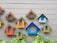 Several colorful, house-shaped frames are mounted on a light-colored wall. The frames vary in colors such as blue, orange, green, and brown. At the bottom of the image, there are green plants growing along the wall.