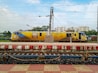 A brightly colored locomotive stands on a railway track, featuring advertisements on its side. The train sits behind a striped platform with a railing and power lines overhead. Buildings are visible in the background under a partly cloudy sky.