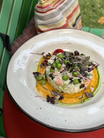 A dish is artfully presented in a white bowl with a variety of fresh ingredients. There's a colorful mix of green peas, microgreens, and possibly diced vegetables on top of a creamy mixture, surrounded by a bright orange puree. The background includes a striped fabric and hints of a vibrant outdoor setting.