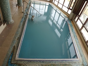 An indoor pool with clear blue water, surrounded by a tiled border and columns. The pool area is enclosed by large glass windows allowing natural light in, and there's a metal handrail leading into the water.