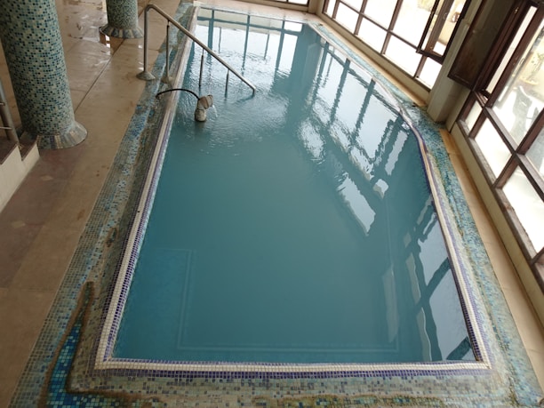 An indoor pool with clear blue water, surrounded by a tiled border and columns. The pool area is enclosed by large glass windows allowing natural light in, and there's a metal handrail leading into the water.