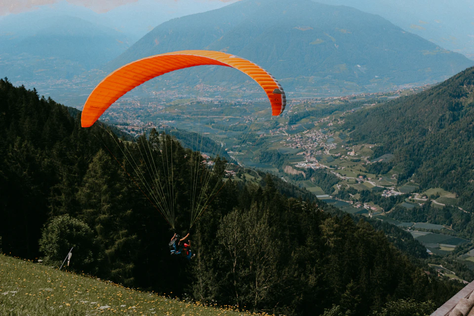 The image depicts a paraglider soaring high above a verdant valley, with an orange parachute billowing behind them against a clear blue sky. The paraglider is positioned centrally in the frame and appears to be flying at a considerable altitude over the lush green landscape below. In the distance, several buildings can be seen nestled among the trees, adding depth to the scene.