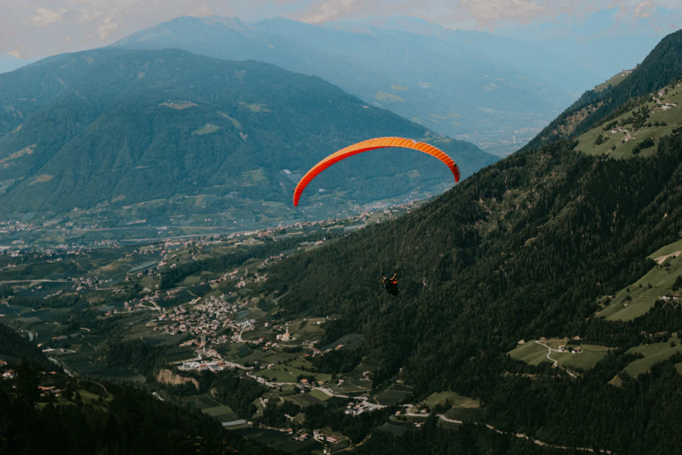 The image depicts a paraglider soaring high above a mountainous landscape, with an orange and white parachute billowing against the backdrop of lush green trees and rolling hills. The paraglider is positioned centrally in the frame, creating a sense of balance between the natural beauty of the surroundings and the exhilarating activity taking place.

The mountainside town below adds depth to the scene, with buildings nestled among the verdant landscape. The sky above is a clear blue, dotted with fluffy white clouds that add to the overall tranquility of the image.