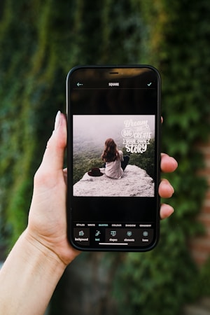 A hand is holding a smartphone displaying an image editing application. On the screen, a picture shows a person with long brown hair sitting on a rock, facing away from the camera, overlooking a misty, forested landscape. The text on the image reads 'Dream big, create your own story'. The phone's screen interface includes several editing options such as styles, write, quotes, color, shadow, and gradient.