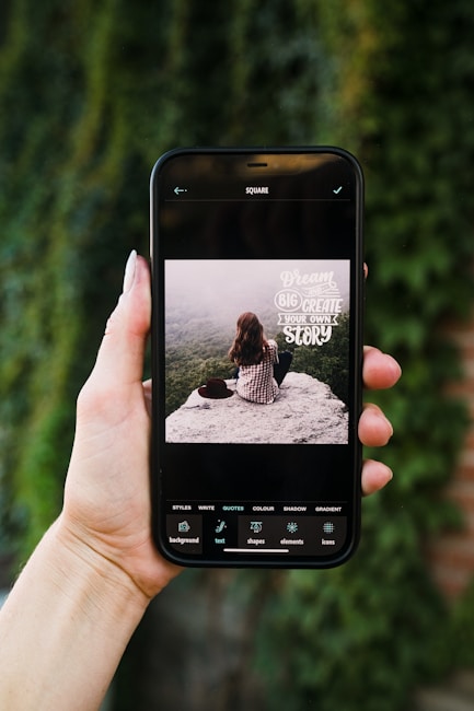 A hand is holding a smartphone displaying an image editing application. On the screen, a picture shows a person with long brown hair sitting on a rock, facing away from the camera, overlooking a misty, forested landscape. The text on the image reads 'Dream big, create your own story'. The phone's screen interface includes several editing options such as styles, write, quotes, color, shadow, and gradient.