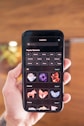 A person holds a smartphone displaying a screen with various digital design elements. The screen shows a search bar at the top followed by a list of 'Popular Elements' categorized into themes such as 3D, Animal, and Arrow. Thumbnails of graphics, including abstract shapes and animal designs, are visible.