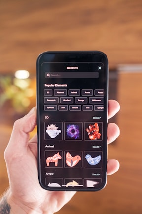 A person holds a smartphone displaying a screen with various digital design elements. The screen shows a search bar at the top followed by a list of 'Popular Elements' categorized into themes such as 3D, Animal, and Arrow. Thumbnails of graphics, including abstract shapes and animal designs, are visible.