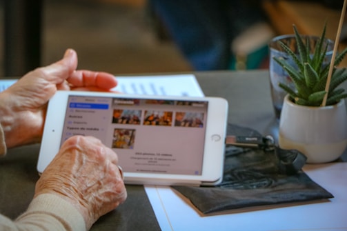An elderly person is holding a tablet, viewing images on the screen. Next to the tablet, there is a potted plant and some papers on the table, creating a cozy and casual atmosphere.