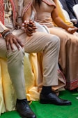 A man and a woman are seated next to each other, holding hands. The man is wearing beige pants, a beige jacket adorned with red patterns, black socks, and black dress shoes. The woman is also dressed in traditional beige attire with red embellishments. Both are accessorized with beaded bracelets.