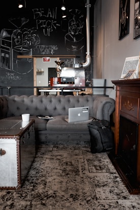 A cozy coffee shop interior features a dark tufted sofa with a laptop and a black backpack placed on it. A metal trunk is used as a coffee table with a white cup on top. Chalkboard art on the wall includes drawings of coffee machines and playful text. The atmosphere is warm and inviting, accentuated by a patterned rug and framed artwork. A roasting machine is visible in the background.