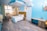 A modern and well-lit hotel room with a double bed featuring white linens, and a wooden headboard with a mustard accent. The room has a wooden floor and is decorated in shades of blue and white, creating a fresh and calming atmosphere. There are two small tables beside the bed, one with a lamp and the other with a phone. A large window with curtains provides a view of a balcony with seating. The room includes a desk and an air conditioning unit on the wall, and there are decorative light fixtures hanging from the ceiling.
