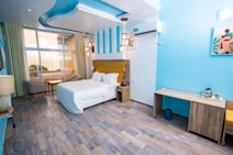 A modern and well-lit hotel room with a double bed featuring white linens, and a wooden headboard with a mustard accent. The room has a wooden floor and is decorated in shades of blue and white, creating a fresh and calming atmosphere. There are two small tables beside the bed, one with a lamp and the other with a phone. A large window with curtains provides a view of a balcony with seating. The room includes a desk and an air conditioning unit on the wall, and there are decorative light fixtures hanging from the ceiling.